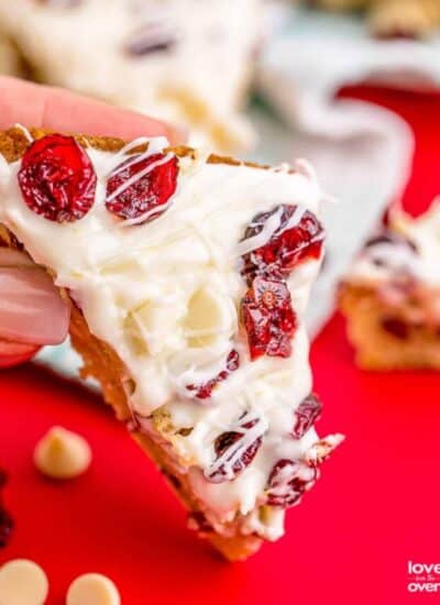 A hand holding a cranberry bliss bar on a red background.