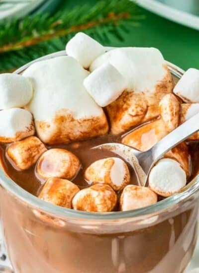 a glass of hot cocoa with marshmallows on top being stirred with a spoon