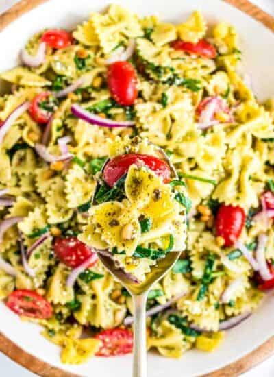 A spoon holding a serving of pesto pasta salad over a bowl full of pasta salad.