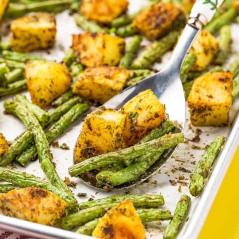 A spoon lifting up green beans and potatoes off a sheet pan