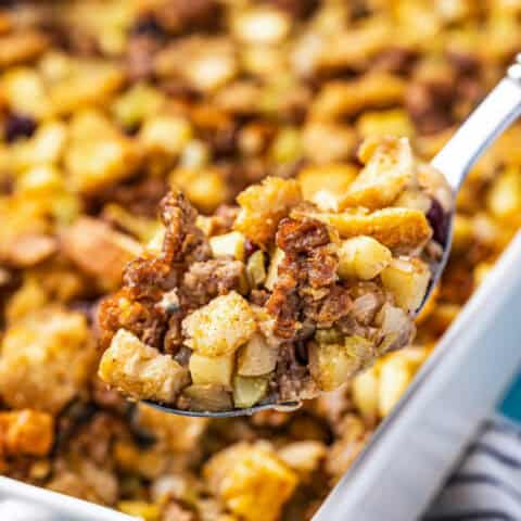 A spoonful of apple sausage stuffing being scooped out of a white casserole dish